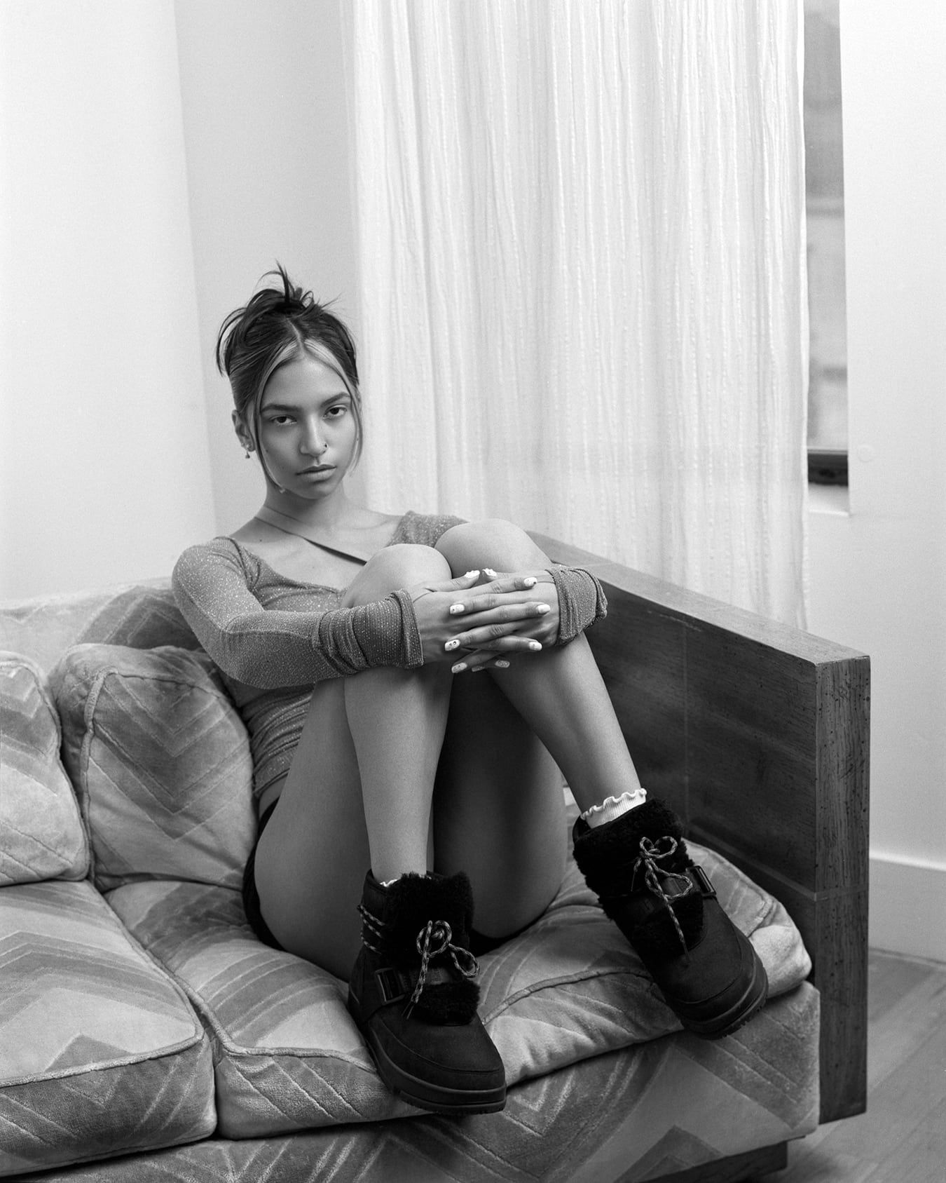 Black and white photo featuring Tara Raani a queer model and actress, know for her role as Zaara Ali on the television sitcom grown-ish. Wearing an oversized sweatshirt, sitting on a 70's sofa, in the background a window with a curtain. Hair in a bun by hairstylist Jason Linkow, photographed by New York fashion photographer Maria Bruun. Photographed using the Mamiya RZ67 camera and Ilford film. Editorial for Flanelle Magazine.