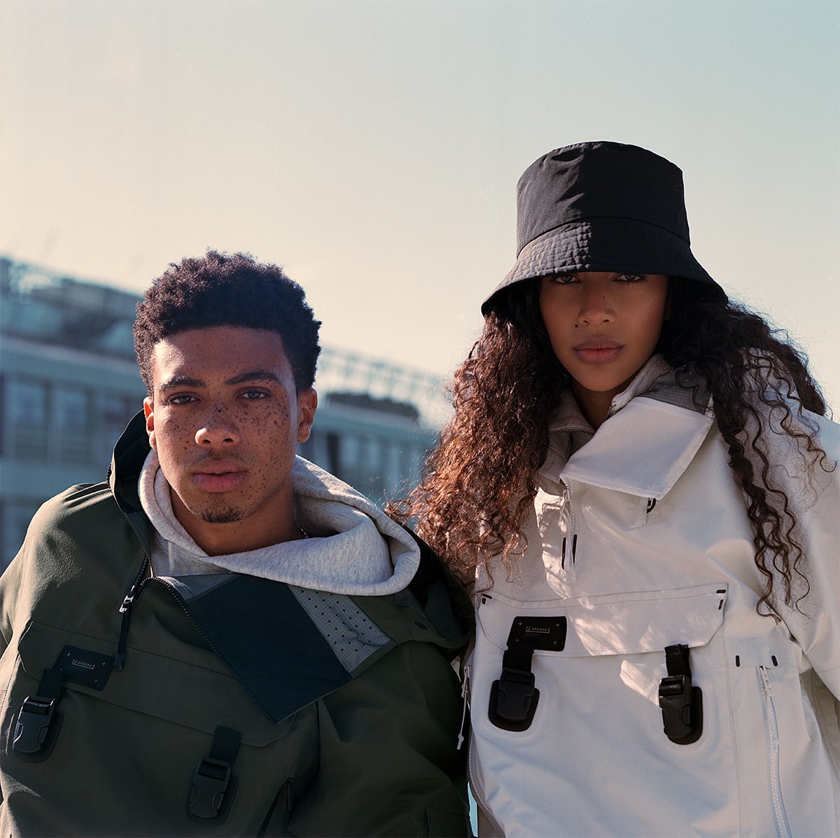 Models Julia Biango and Leron Nurse wearing Armada ski outerwear jackets, her jacket is white his jacket is army green. She is wearing a black bucket hat. They are standing in front of an industrial building it is a sunny day, blue sky. The male model has a lot of freckles. Look-book photographed by New York photographer Maria Bruun. The image was taken using the Hasselblad 500CM camera and Kodak Portra film.