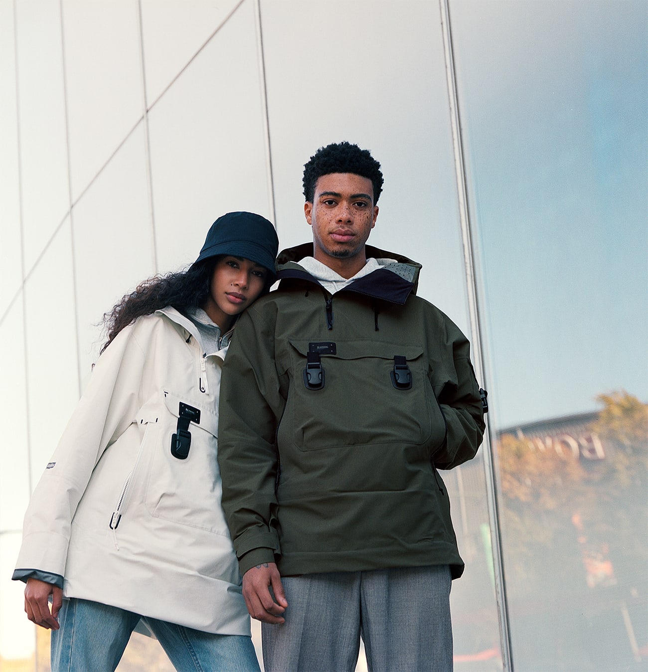 Models Julia Biango and Leron Nurse wearing Armada ski outerwear jackets, her jacket is white his jacket is army green. She is wearing a black bucket hat. They are standing in front of an industrial Manhattan building it is a sunny day, blue sky. The male model has a lot of freckles. Look-book photographed by New York photographer Maria Bruun. The image was taken using the Hasselblad 500CM camera and Kodak Portra film.