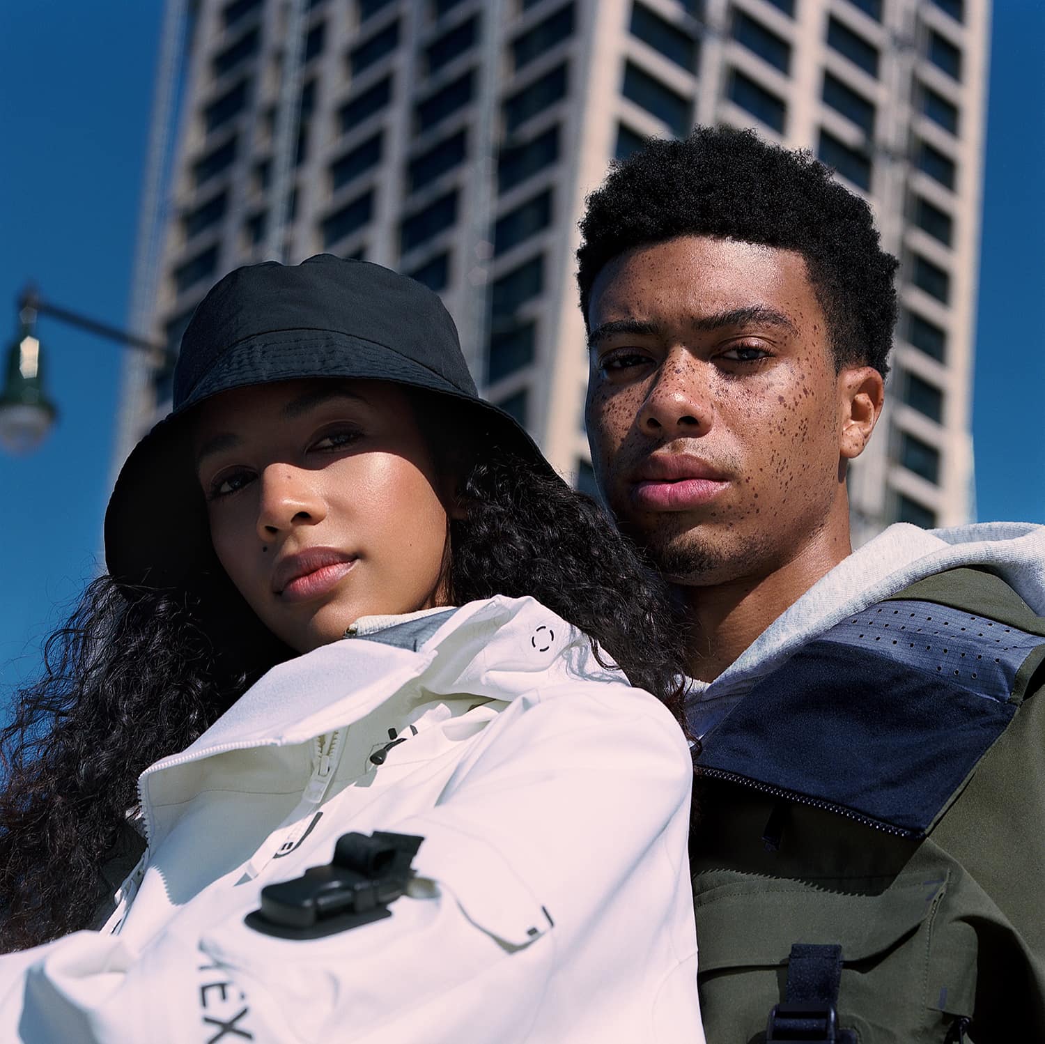 Julia Biango and Leron Nurse wearing Armada ski outerwear jackets, her jacket is white his jacket is army green. She is wearing a bucket hat. The sky is blue and in the background a tall skyscraper building and lamppost. The male model has a lot of freckles. Lookbook photographed by New York photographer Maria Bruun. The image was taken using the Hasselblad 500CM camera and Kodak Portra film.