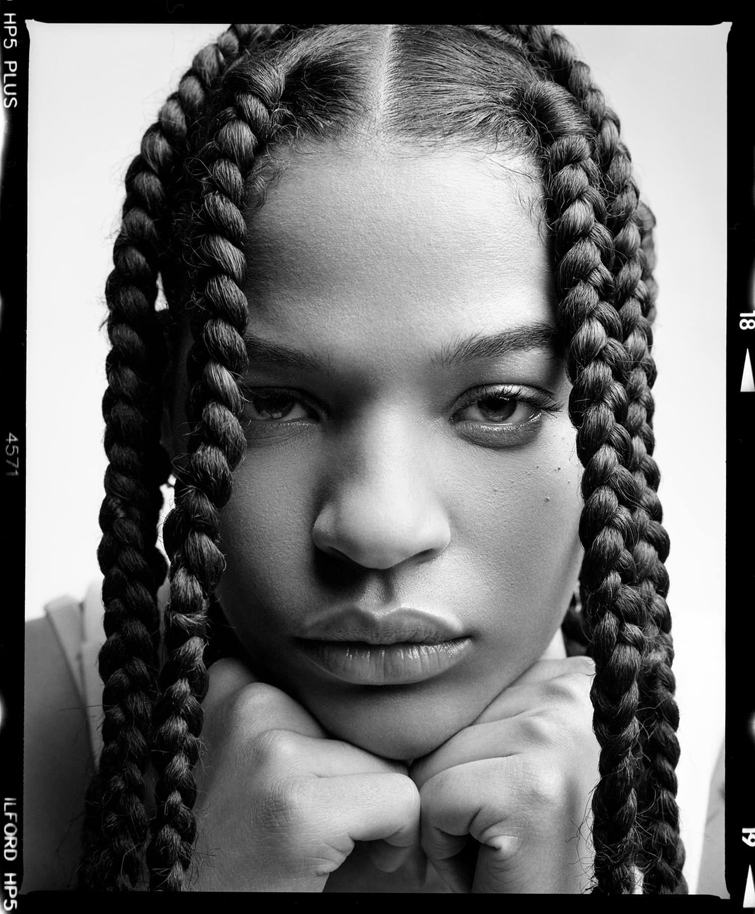 Female Spirit model Candace Demers, sitting with er face in her hands, looking into the camera. Long braids. Portrait photographed by New York photographer Maria Bruun, studio photography setup, photographed using a Mamiya RZ67 camera and Ilford HP5 black and white film.
