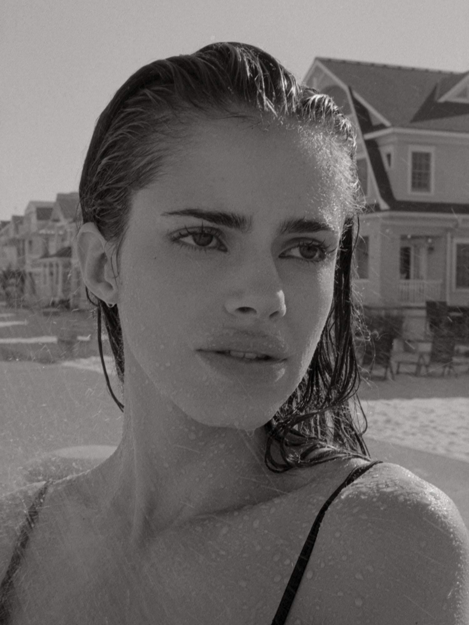 Black and white photo of female model Carmin Valencia on a New Jersey beach, her hair is wet, water droplets on her face and body, water splashing on her face, she is wearing a black Zara dress, vacation houses in the background. Photographed by New York photographer Maria Bruun. Nikon film camera and Ilford film. "High Tide" Fashion editorial for Nasty Magazine.