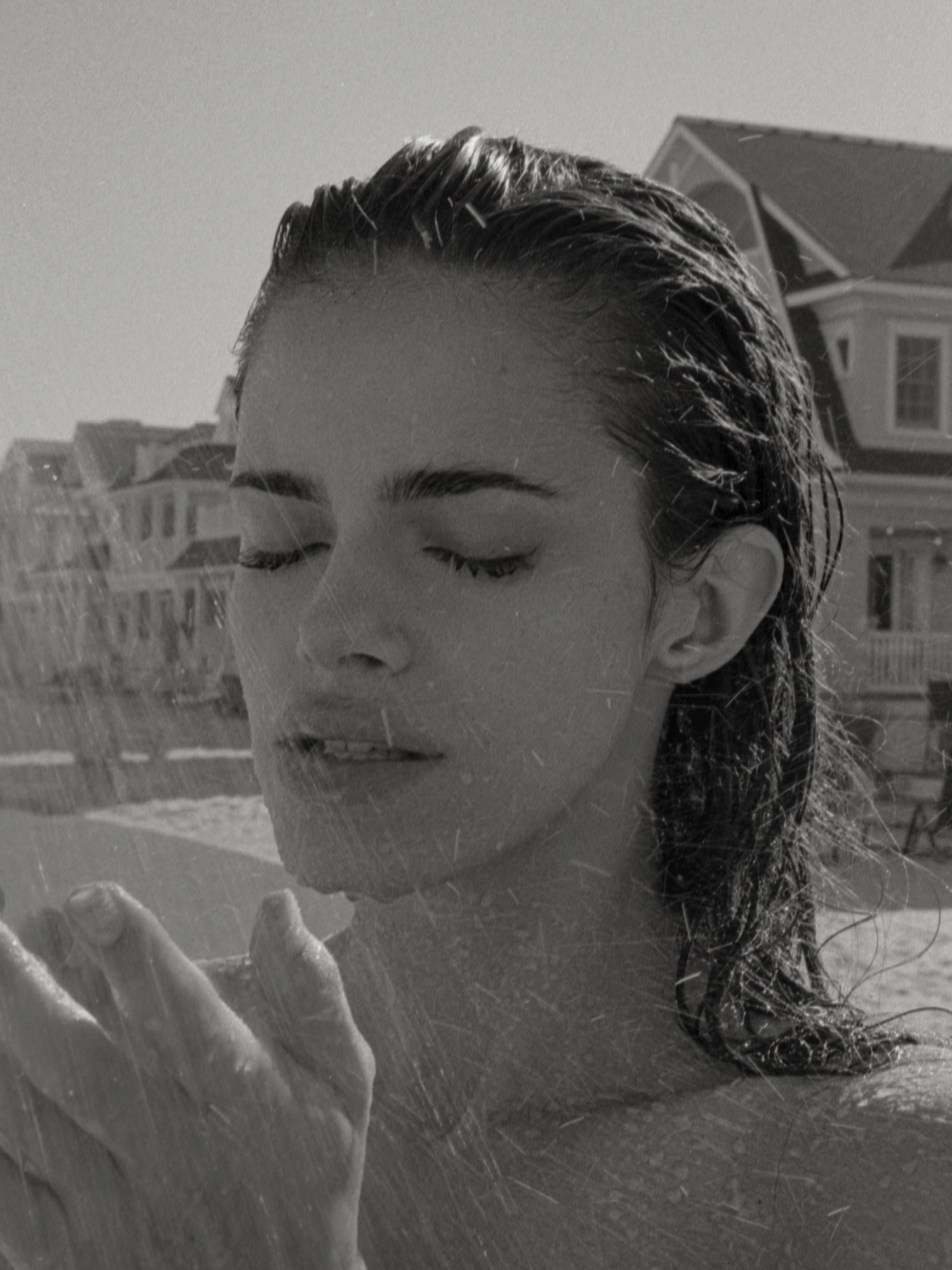 Black and white photo of female model Carmin Valencia on a New Jersey beach, her hair is wet, water droplets on her face and body, water splashing on her face, vacation houses in the background. Photographed by New York photographer Maria Bruun. Nikon film camera and Ilfordfilm. "High Tide" Fashion editorial for Nasty Magazine.