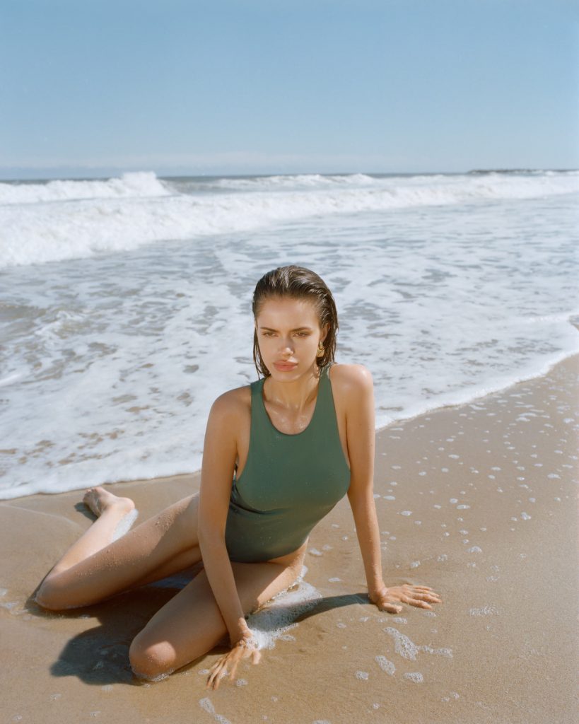 Female model Carmin Valencia wearing a green Zara bathing suit sitting in the sand on a New Jersey beach, in the back ground there is blue sea and white, crashing waves. her hair is wet and she is sun tanned. Photographed by New York photographer Maria Bruun. Mamiya RZ67 camera and Kodak Portra film. 