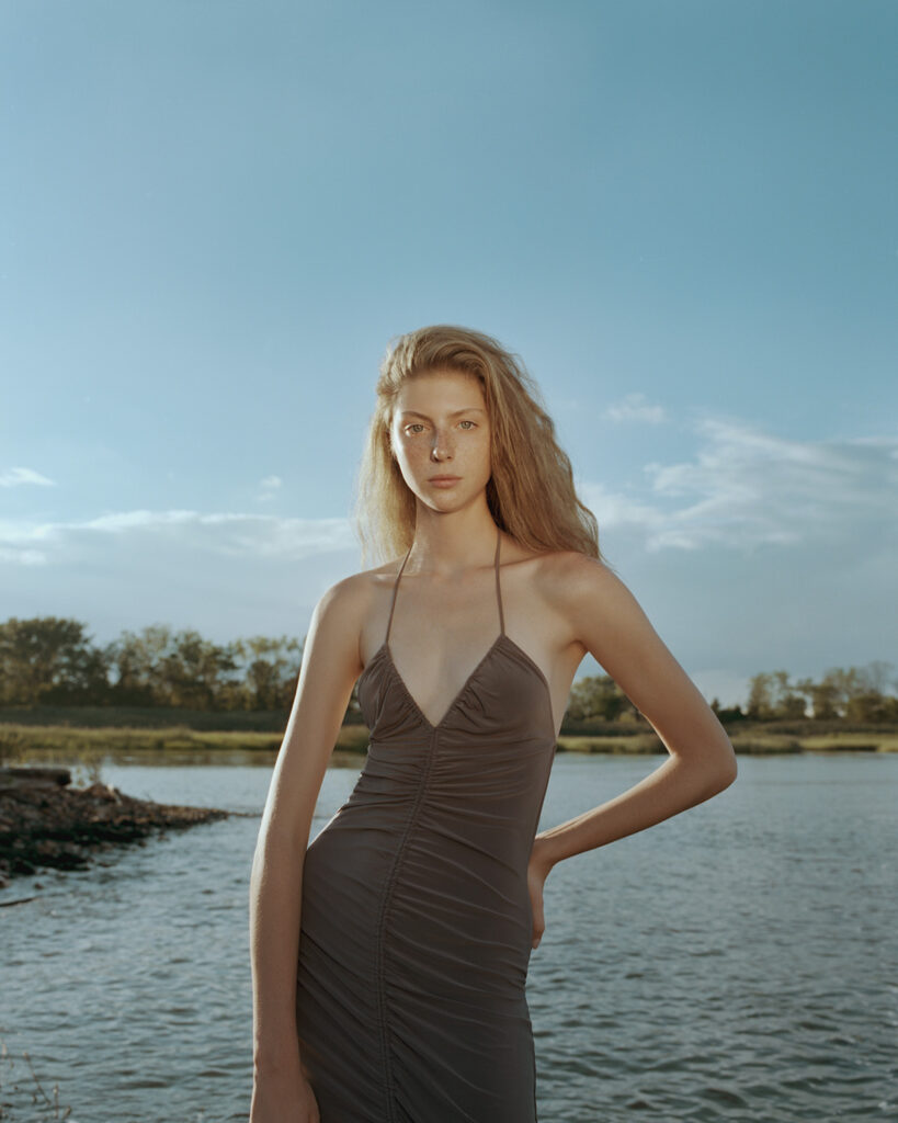 Ford model Polina Sharpran, standing by the Hudson River looking into the camera, bright green eyes and freckles, wearing a brown Zara dress, her hand on her hip. Trees, water and nature in the background. A bright sunny day.