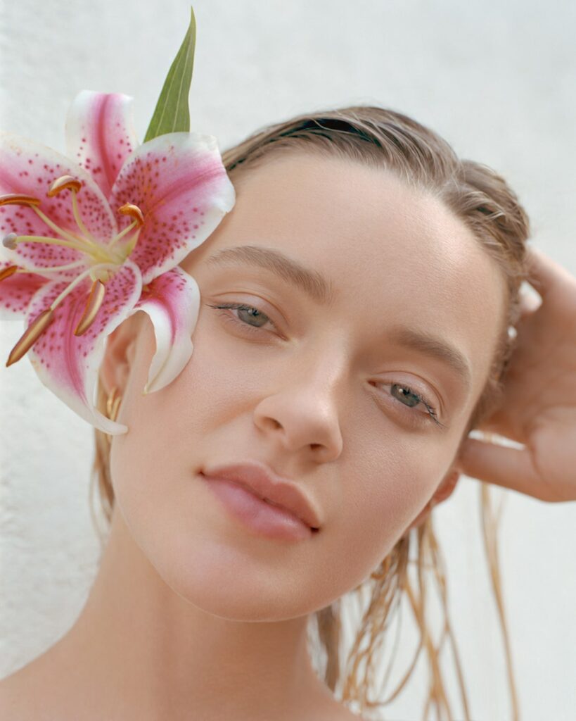 Color portrait of female AMP model Sam Romberger, she is wearing a pink flower in her blonde hair, her head is tilted and her hair is wet. She has a soft smile and light green eyes. Location Jersey City. Editorial for Solstice Magazine. Photographed by New York photographer Maria Bruun. Shot on film with the Mamiya RZ67 camera and Kodak Portra Film.