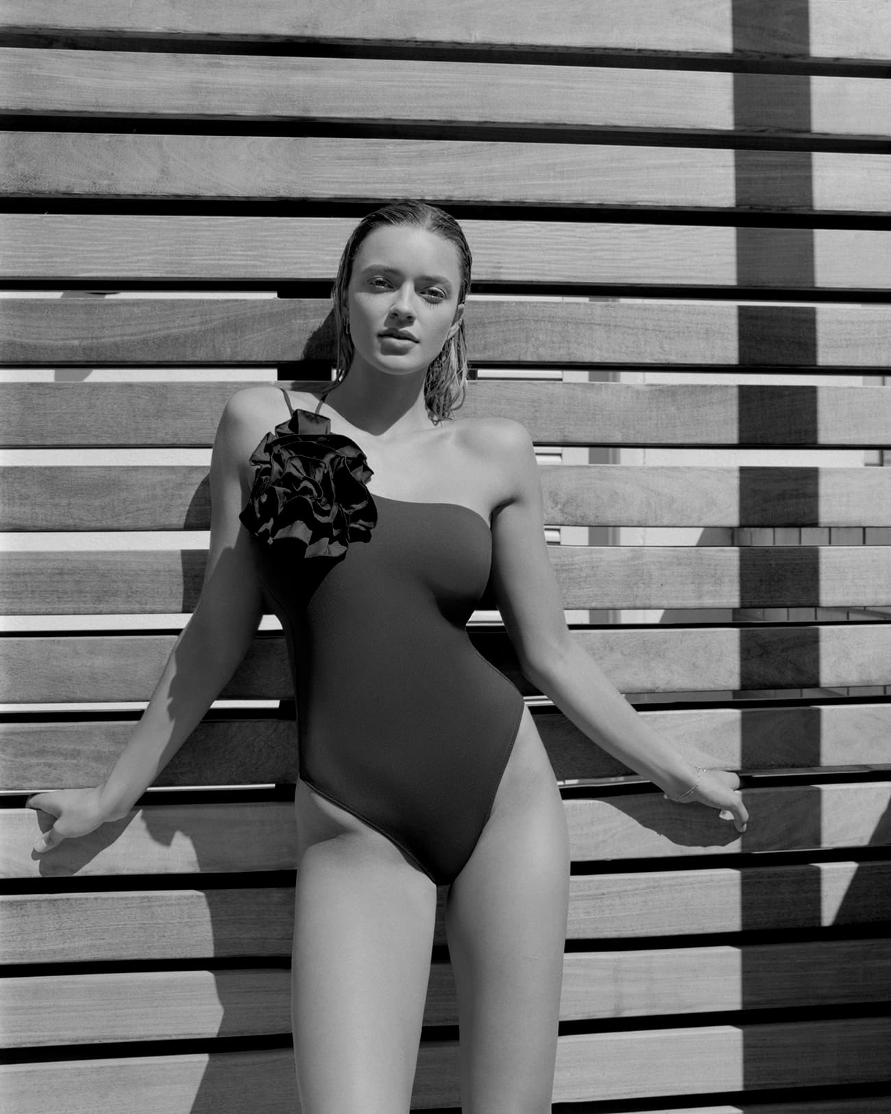 Female APM model Samantha Romberger is wearing a Zara bathing suit with a flower, her hair is wet and combed back. She is standing against a wooden fence. Photographed by New York photographer Maria Bruun. Photographed using a Mamiya RZ67 camera and Ilford black and white film. Editorial for Solstice Magazine.