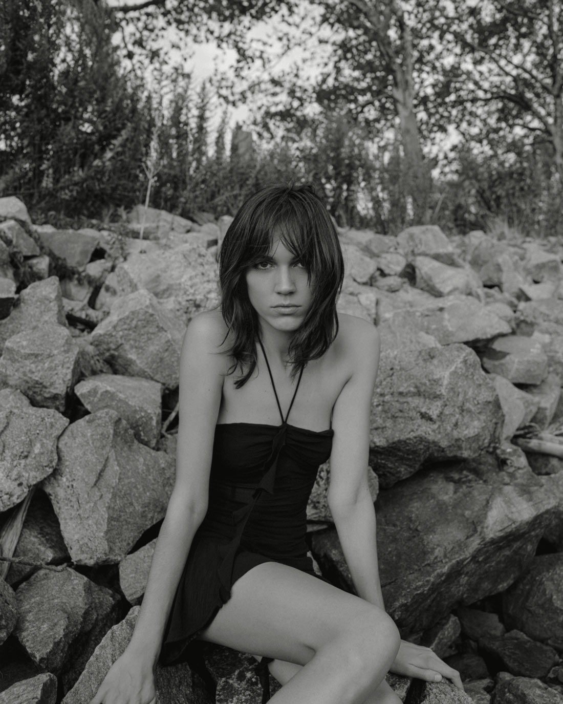 Female model Abby Blaine is sitting on rocks, trees in the background, she is wearing a Zara dress, her hair is down, she has bangs. Photographed by New York photographer Maria Bruun, photographed using a Mamiya RZ67 camera and Ilford black and white film.