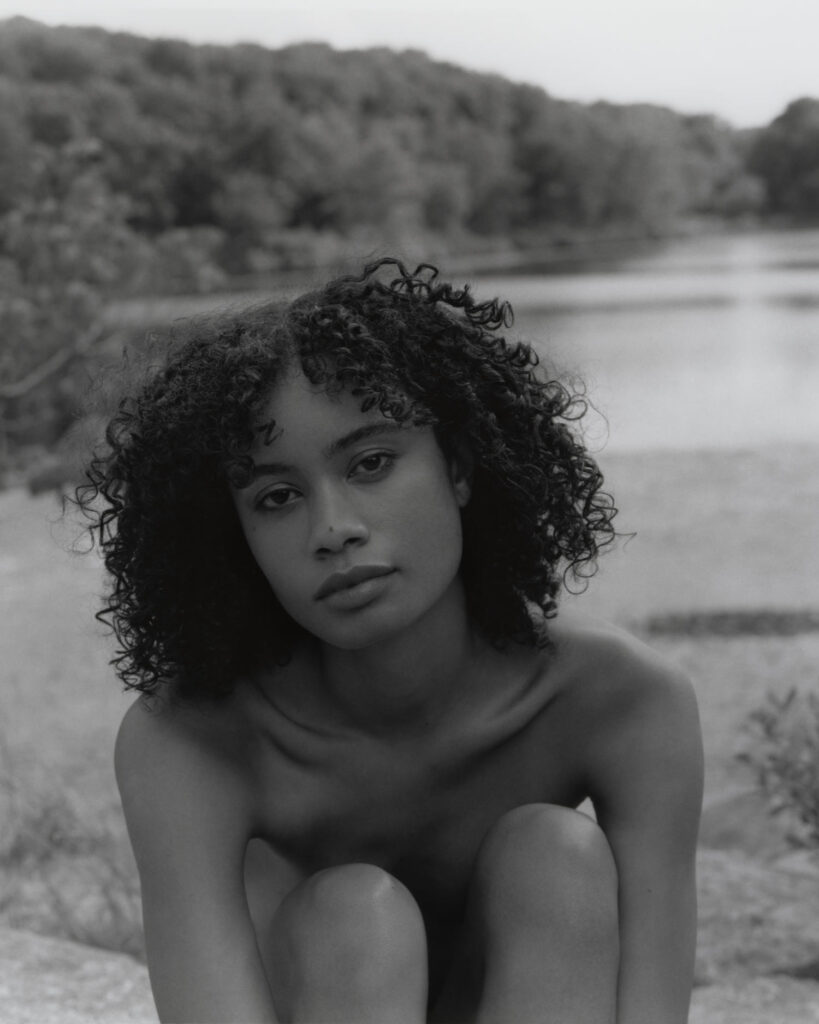 Black and white photo of female model Destene Kinser. Sitting on rocks in Harriman State Park in front of a lake, the forest in the background. She has dark curly hair, her arms wrapped around her legs looking into the camera. Editorial for Cake Magazine photographed by New York photographer Maria Bruun.